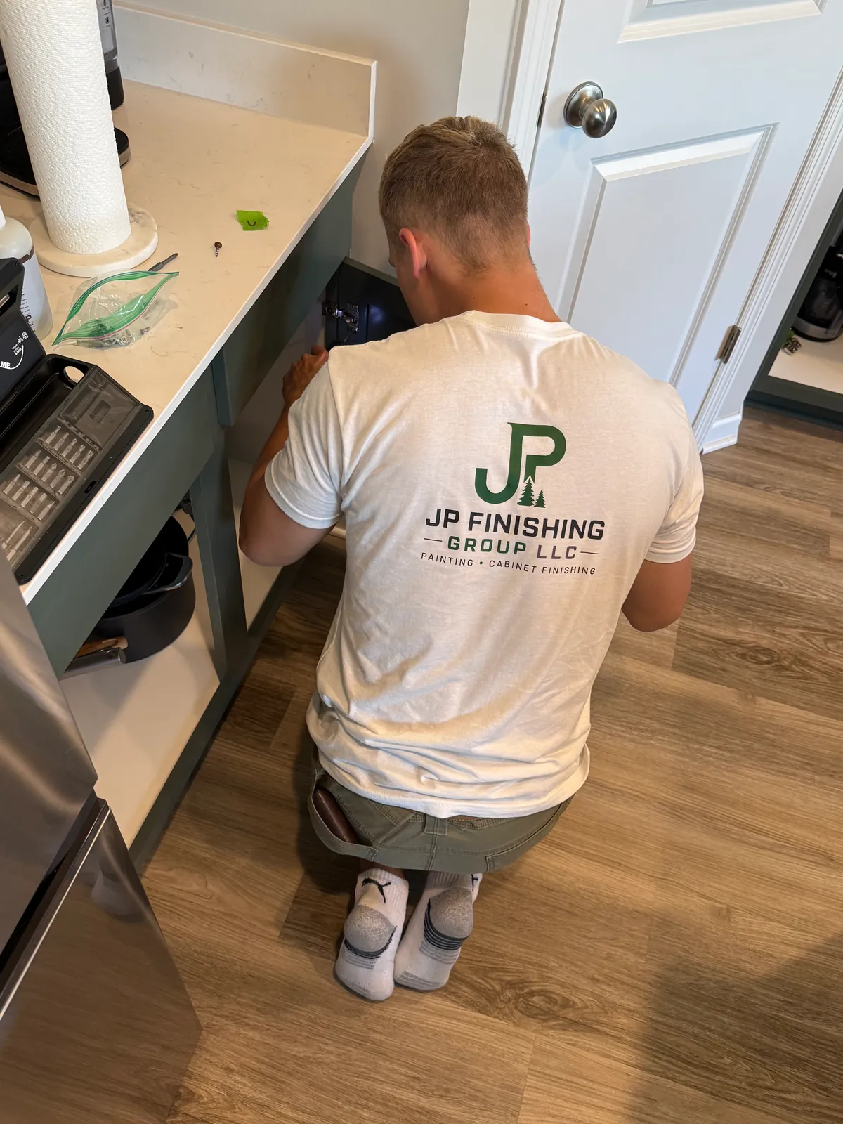 A JP Finishing Group crew member in a company T-shirt kneeling to adjust a Pewter Green base cabinet in a St. Michael kitchen.